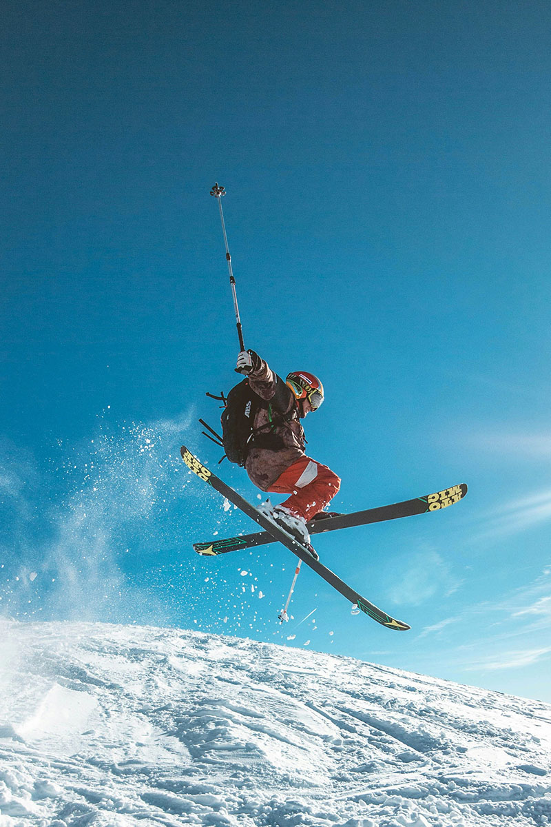 Skifahrer in roter Hose und schwarzer Jacke führt einen Sprung auf Skiern aus, mit Schnee und blauem Himmel im Hintergrund.