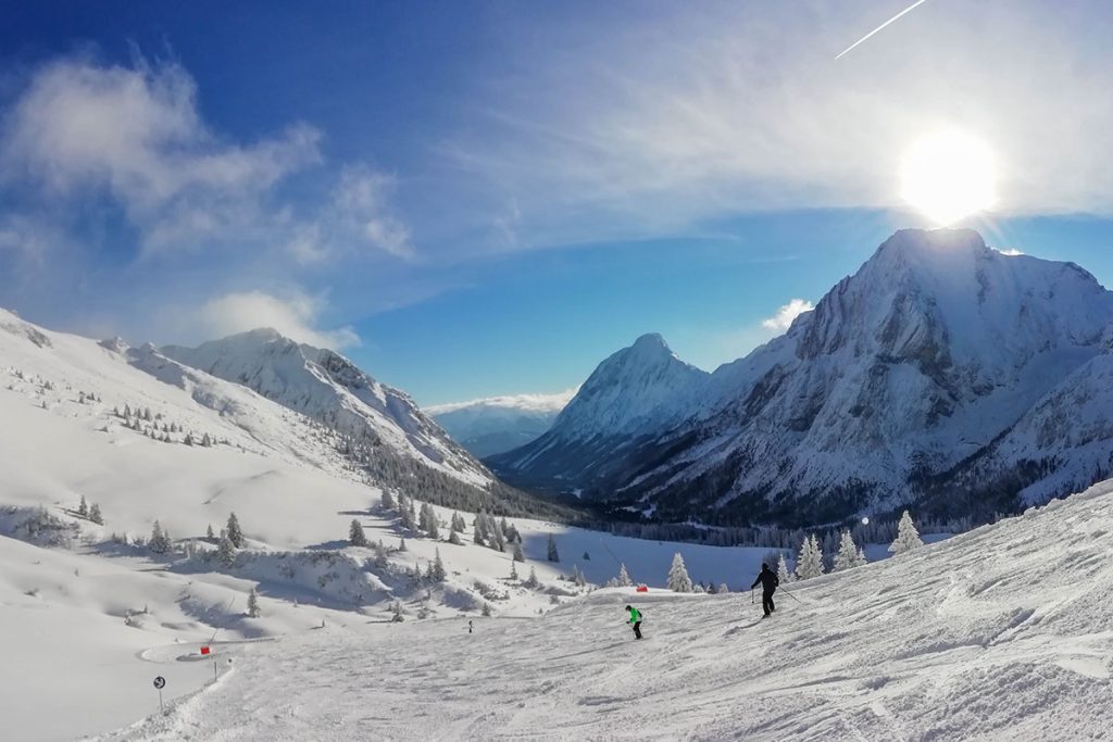 Skifahrer genießen eine sonnige Piste inmitten verschneiter Berge und Tannenwälder, mit majestätischen Gipfeln im Hintergrund.