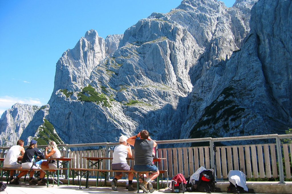 Wanderer genießen den Wilden Kaiser und die Aussicht auf steile Felswände und grüne Berghänge von einer Terrasse aus, mit klarem Himmel im Hintergrund.
