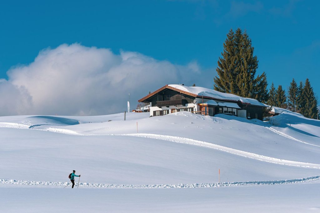 Langläufer in verschneiter Landschaft vor einem traditionellen Chalet mit Blick auf schneebedeckte Hügel und klarem blauen Himmel.