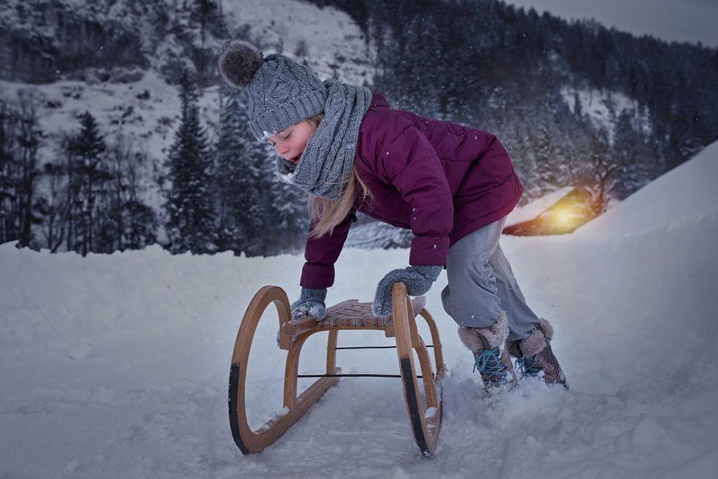 Kind beim Schlittenfahren in der Dämmerung einer winterlichen Landschaft, warm eingepackt mit Mütze und Schal, umgeben von Schnee und Bergen.